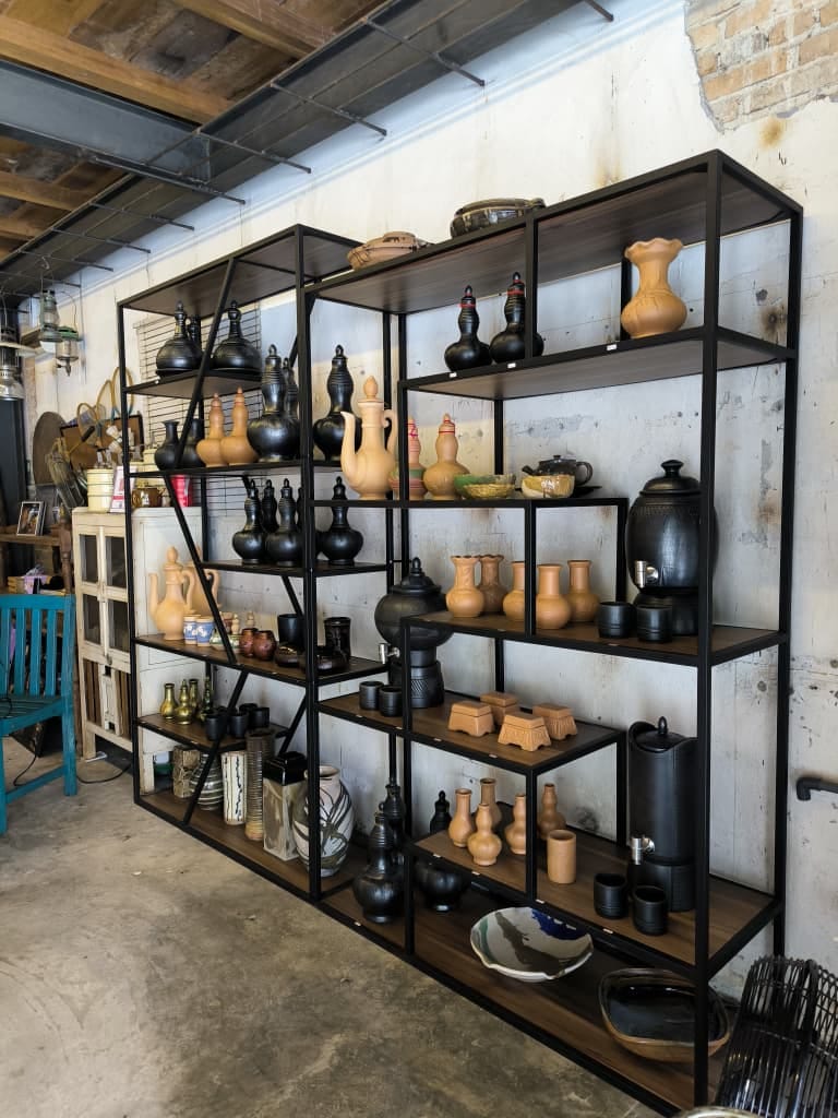 A wall of finished ceramics on steel shelving — black-fired Labu Sayong gourds, terracotta jugs, cups and bowls — in a stockist's display room