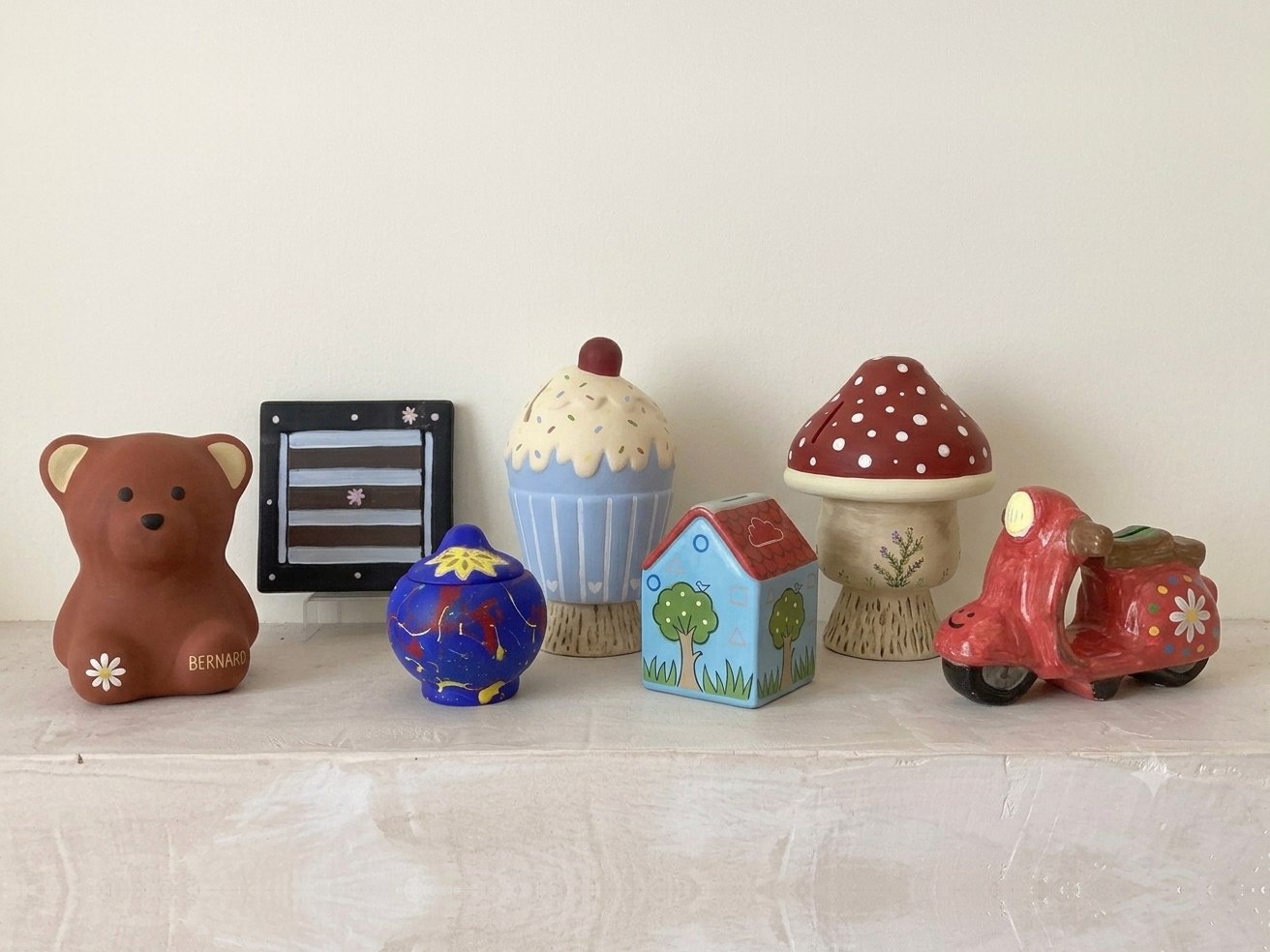 Seven hand-painted ceramic pieces on a shelf — a bear coin bank, a painted tile, a blue lidded pot, a cupcake jar, a house, a toadstool and a red scooter